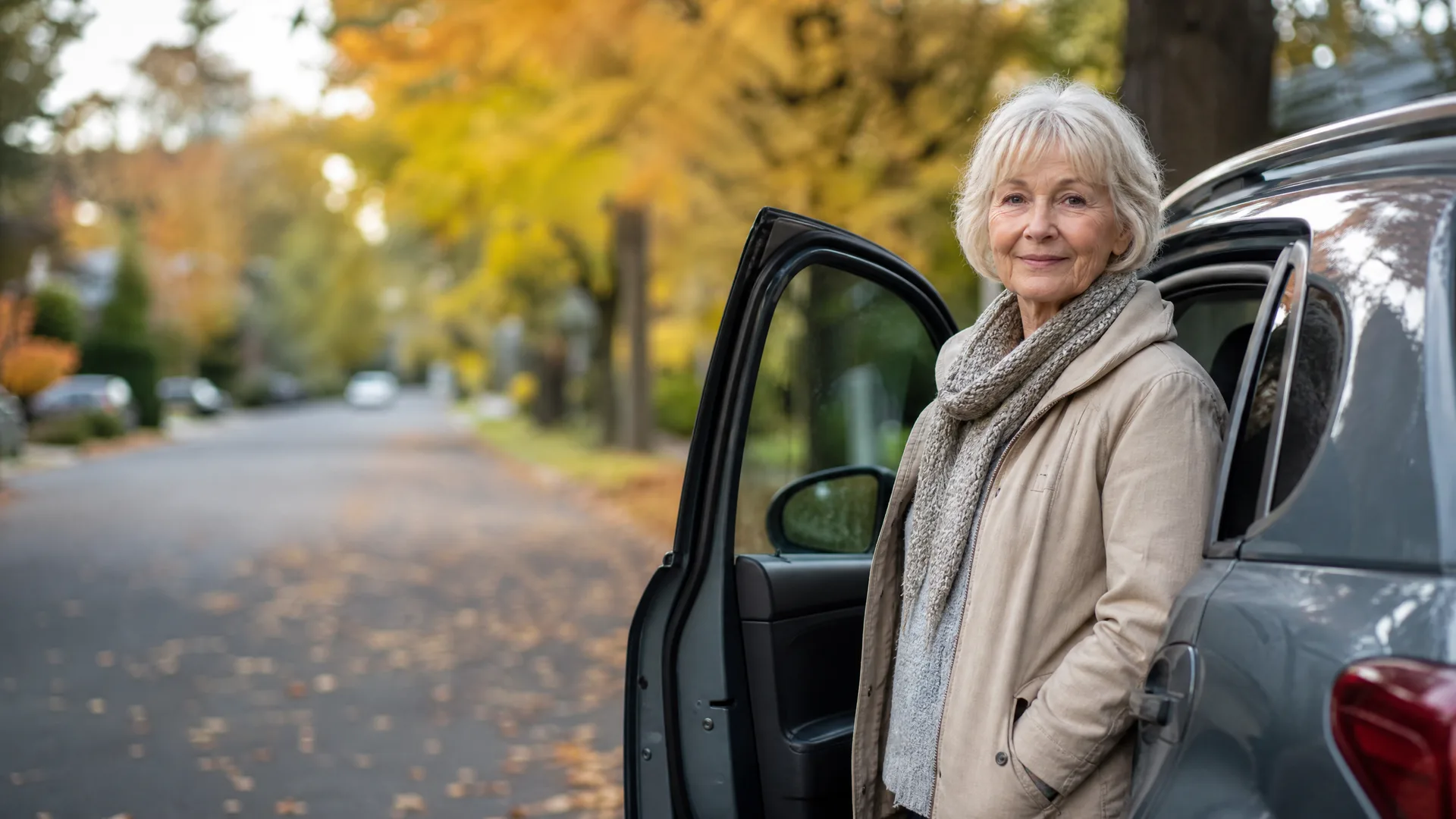 An older woman with white hair, wearing a beige jacket and knit scarf, stands beside an open rear door of a gray car on a tree-lined residential street with yellow autumn foliage.