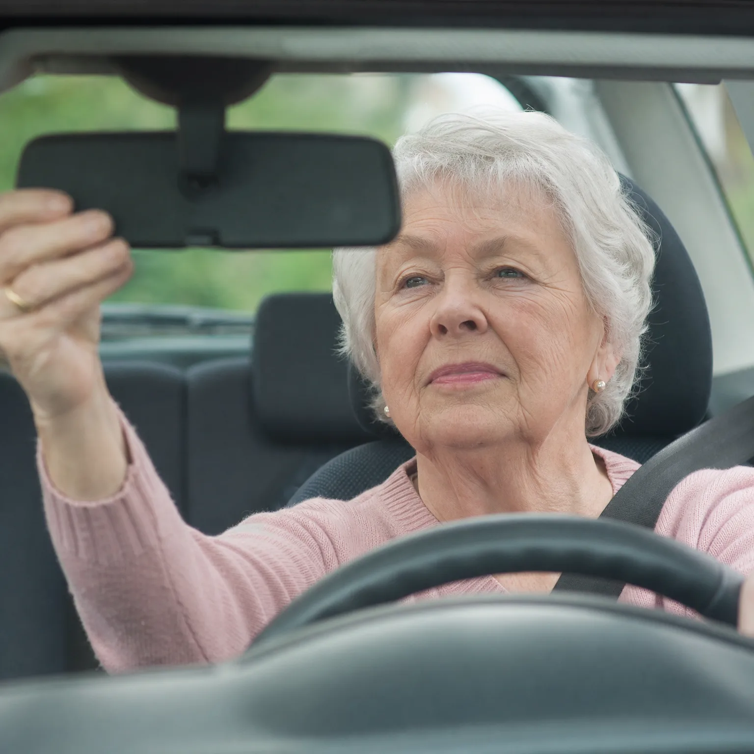 An older woman with short white hair in a pink sweater sits in a car's driver seat, reaching up with her left hand to adjust the rearview mirror, with green foliage visible through the windows.
