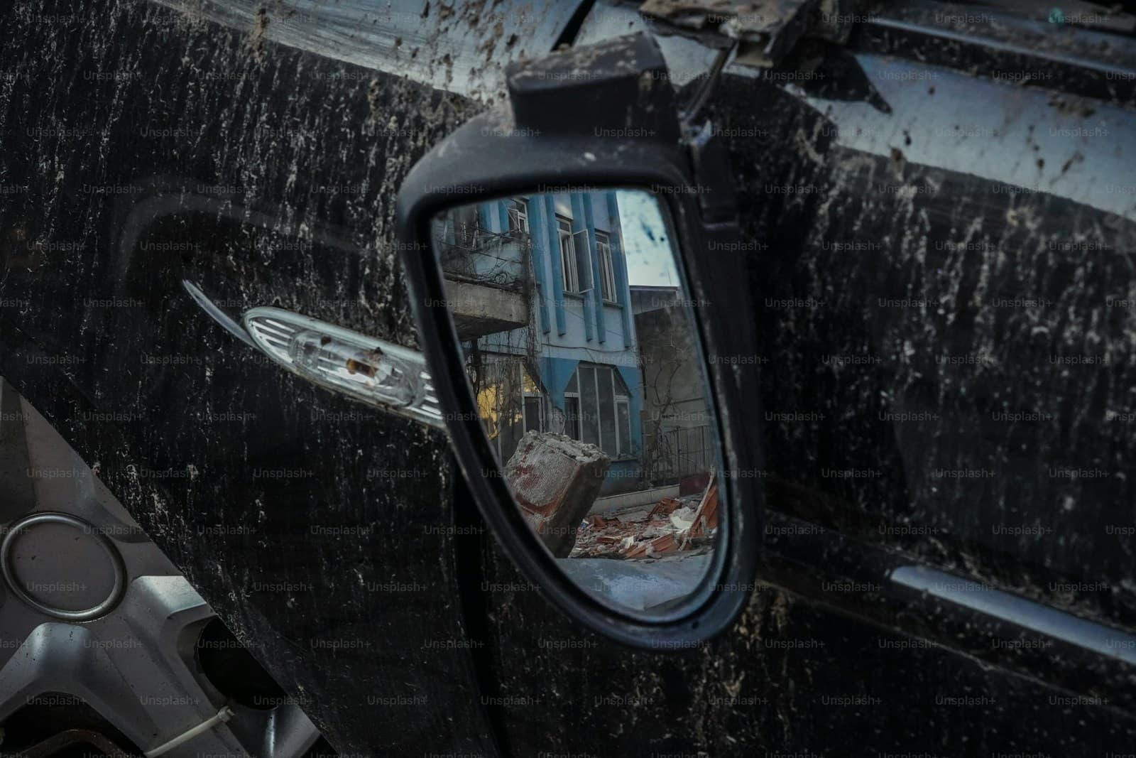 Vehicle side mirror reflecting a blue-windowed building, mounted on dark wet car surface