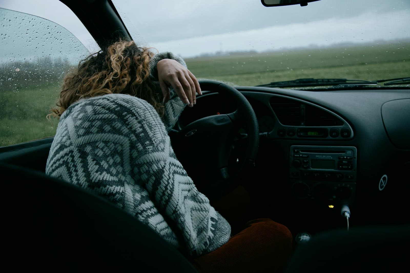 Driver in a patterned sweater slumped over the steering wheel with rain on the windshield